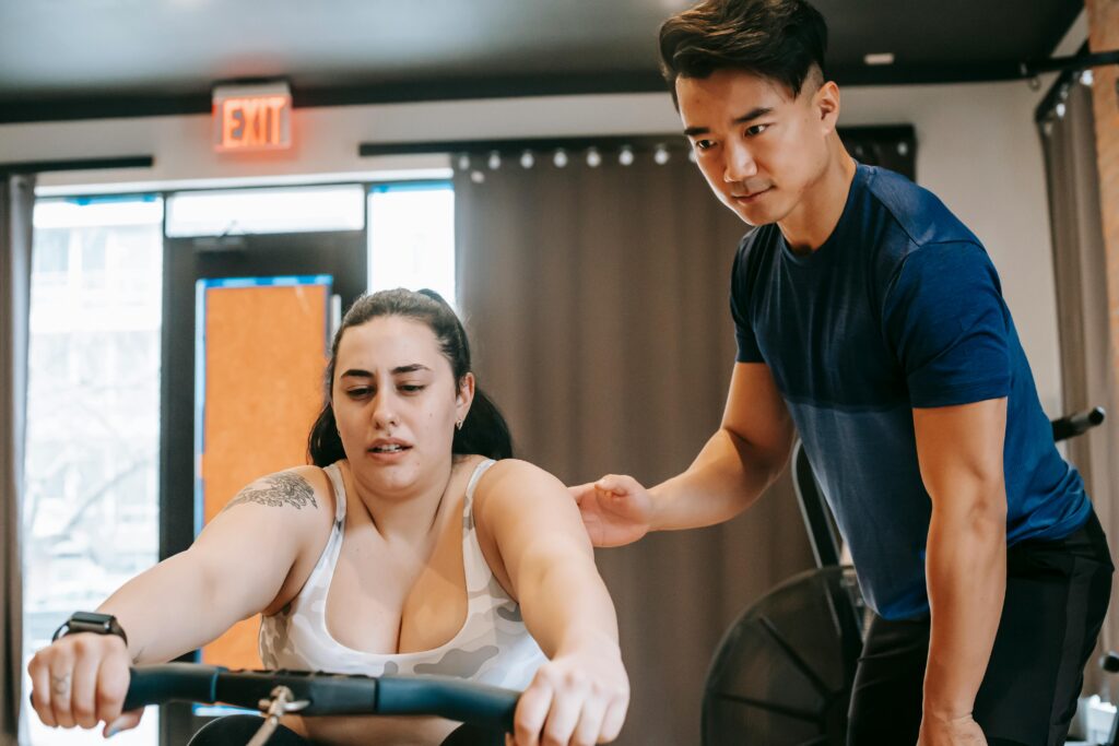 Young handsome ethnic experienced trainer supporting plus size woman working out on rowing machine in gym